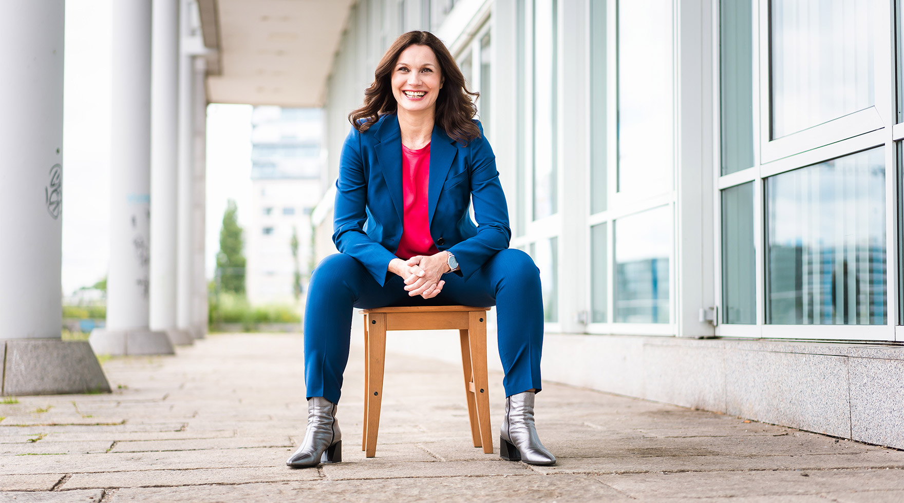 Business Coach Alice Fiedler sitzt auf einem Holzhocker vor einem Firmengebäude und lacht