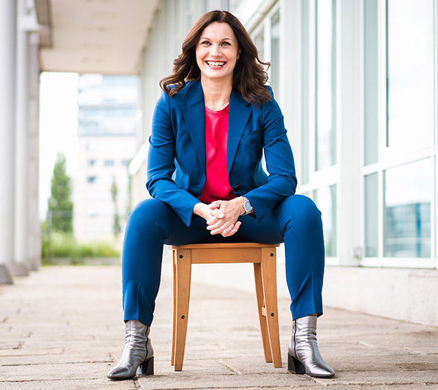 Business Coach Alice Fiedler sitzt auf einem Hocker vor dem Firmengebäude. Sie trägt einen blauen Anzug, ein pinkfarbenes Shirt und silberne Stiefel.