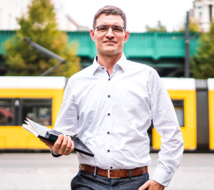 Inhaber von phonux events läuft auf de Straße mit einem Notizbuch und Handy in der Hand. Im Hintergrund fährt eine Straßenbahn vorbei.